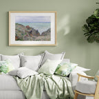 A framed landscape art print depicting a coastal cliffside with green hills and a blue sea, hung on a sage green wall above a modern white sofa styled with green and white pillows and a light green throw blanket.