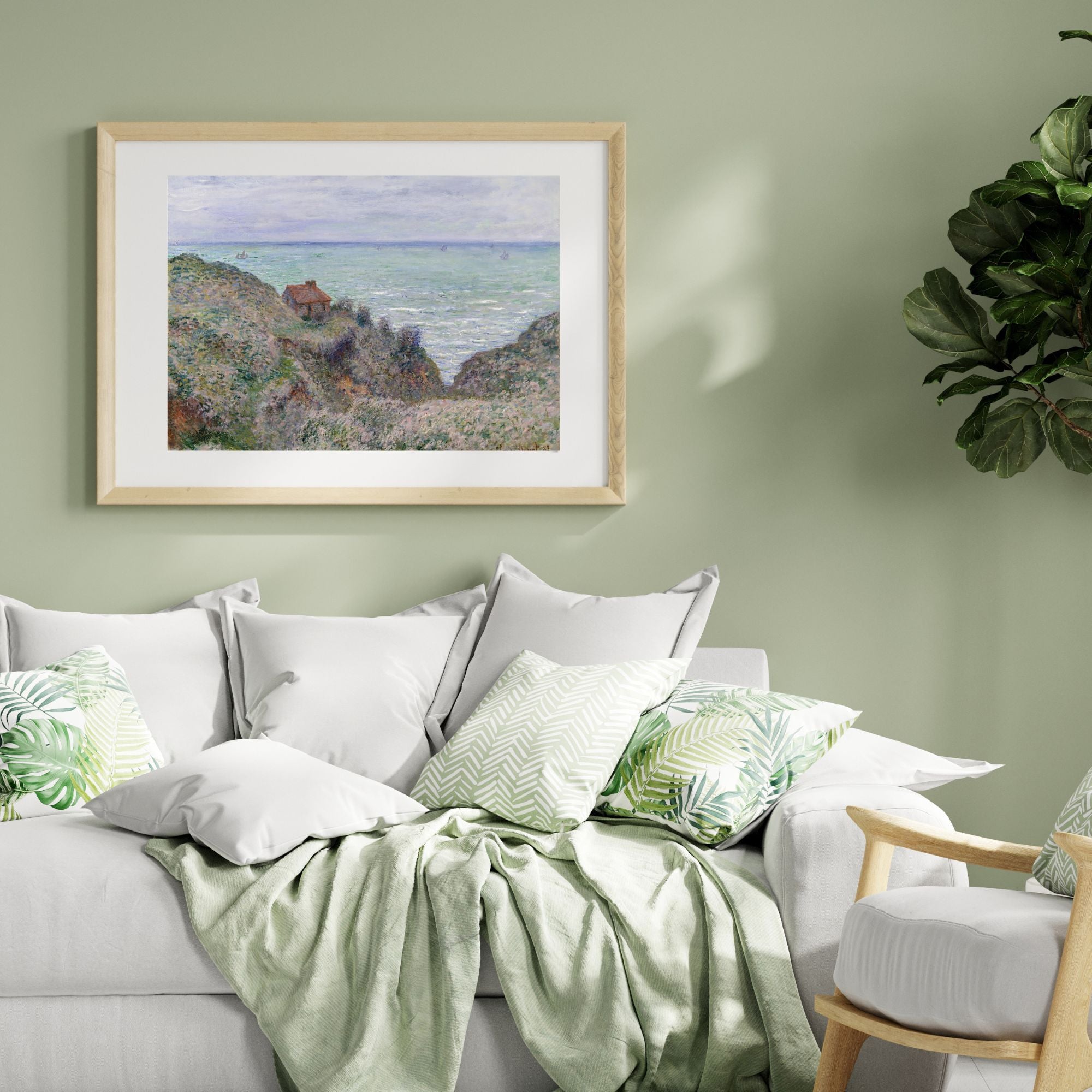 A framed landscape art print depicting a coastal cliffside with green hills and a blue sea, hung on a sage green wall above a modern white sofa styled with green and white pillows and a light green throw blanket.